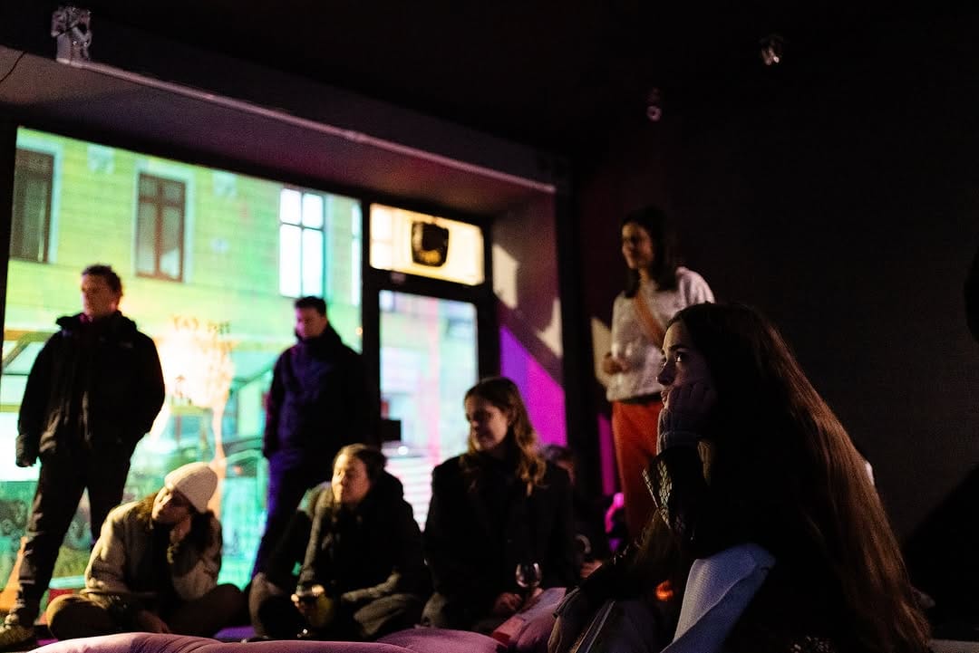 A group of people, some sitting, some standing, watching a three-channel video in a black exhibition space.