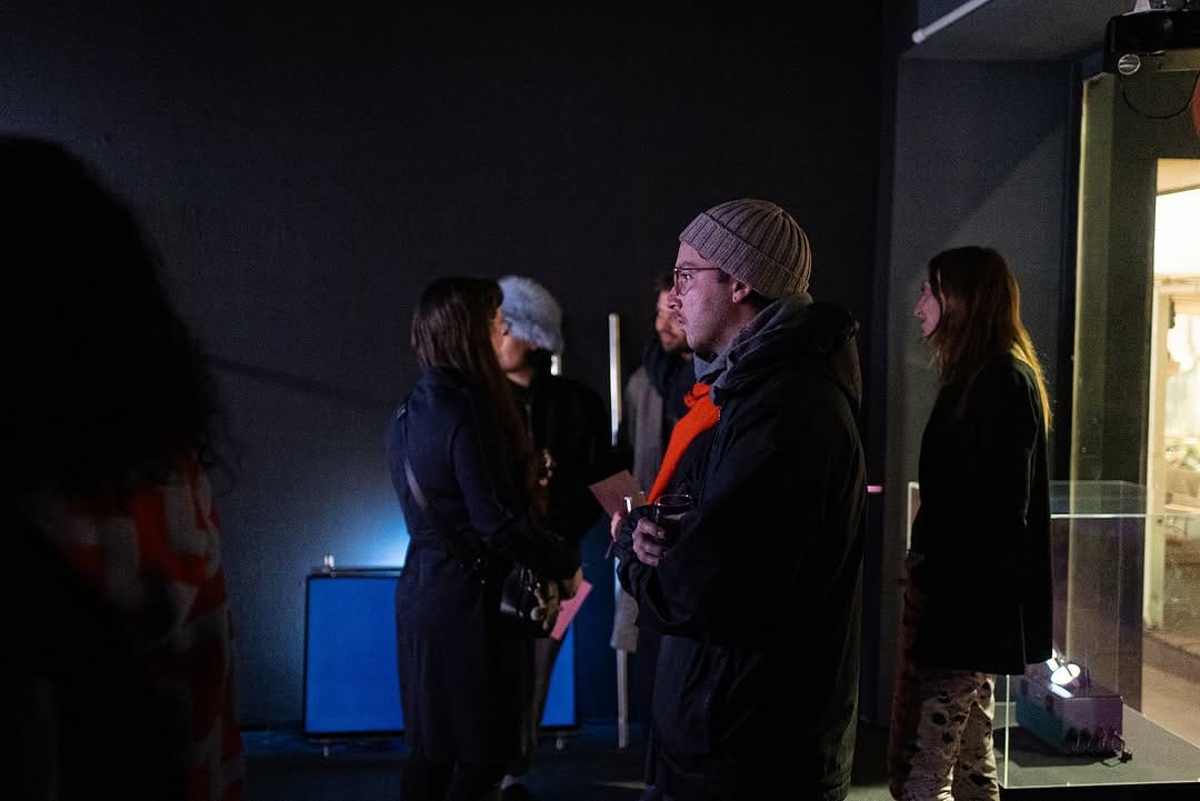 A group of people standing and watching a three-channel video in a black exhibition space. TX-1 is on the right.