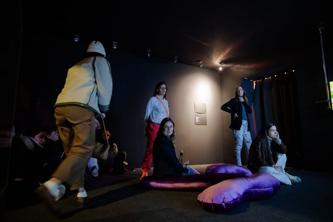 People standing and sitting watching a three-channel video in a black gallery space. They are lit by the TV glow. Some sit on purple organically-shaped velvet-covered cushions.