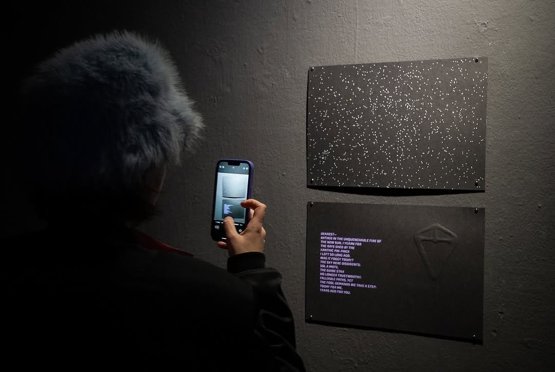 A person taking a photo of my prints of the night sky of Proxima Centauri B. A poem is on the bottom print: DEAREST—//BATHED IN THE UNQUENCHABLE FIRE OF//THE NEW SUN, I YEARN FOR//THE RAYS SHED BY//THE XANTHIC PIN-PRICK//I LEFT SO LONG AGO.//WAS IT FOGGY TODAY?//THE SKY HERE DISORIENTS://SOL A MOTE,//THE GUIDE STAR//NO LONGER TRUSTWORTHY.//FALLIABLE PATHS,//YET THE FOOL DEMANDS WE TAKE A STEP://TODAY FOR ME,//YEARS AGO FOR YOU.