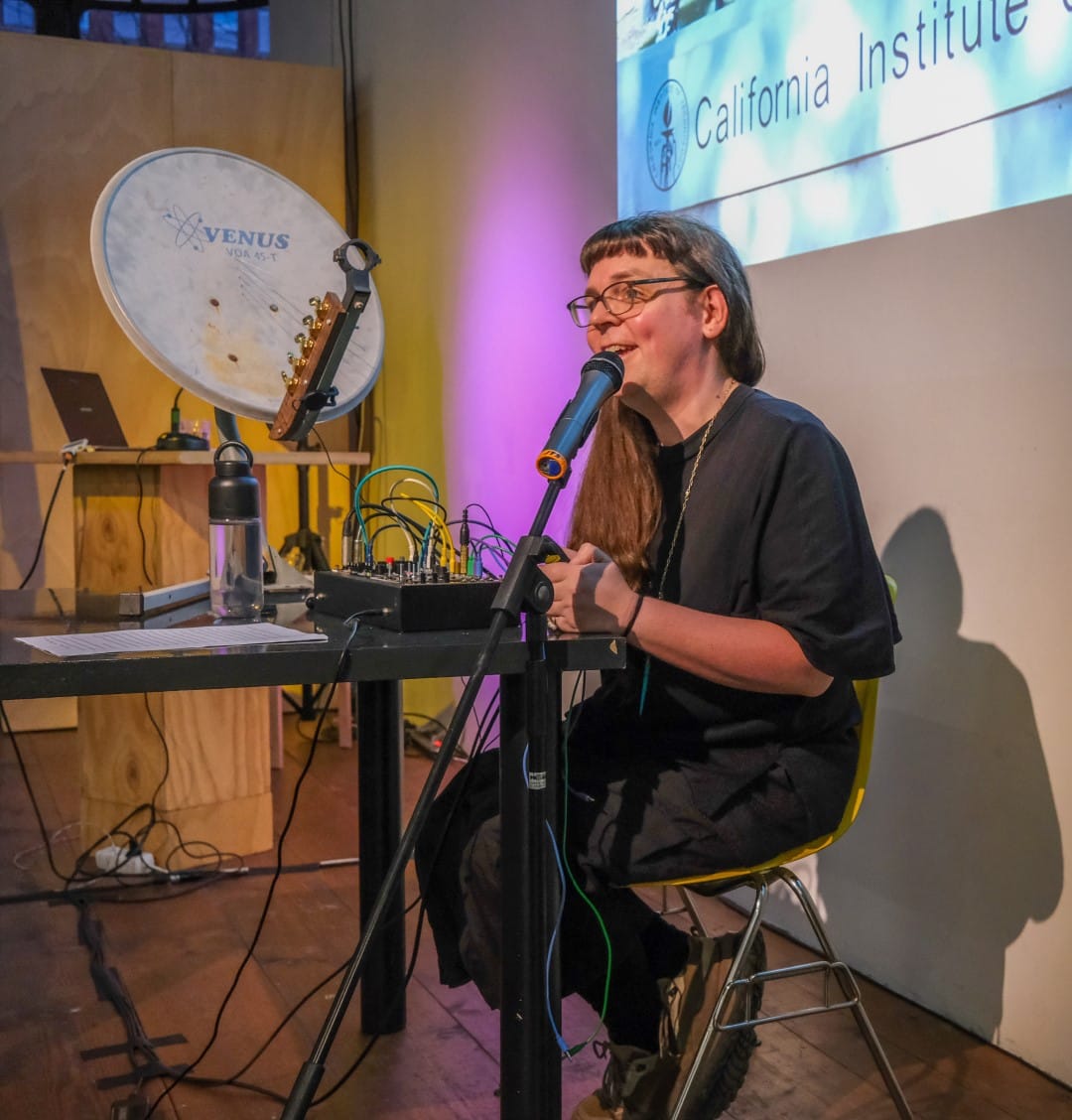 Adriana Knouf sitting down and speaking about Caltech during her lecture-performance. She sits in front of a satellite dish instrument and a small modular synthesizer system.