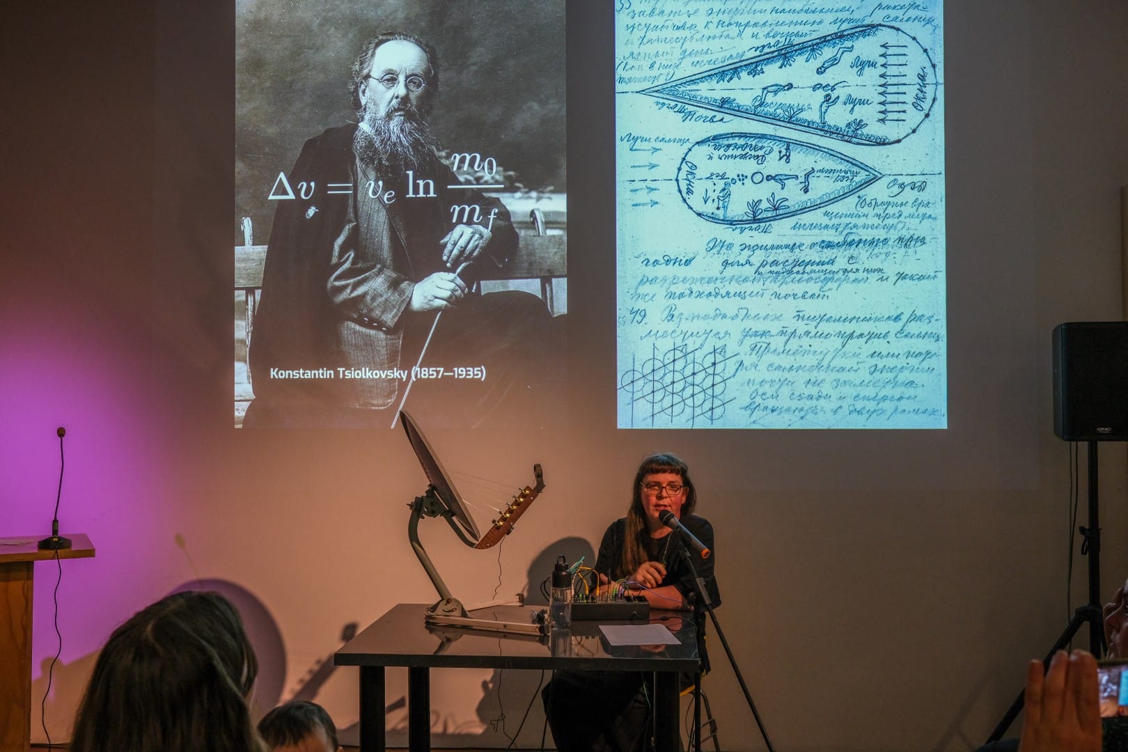 Adriana Knouf during her lecture-performance, with an image of Konstantin Tsiokovsky and his rocket equation projected behind her. She sits down at a table with her satellite dish stringed instrument and a small modular synthesizer system.