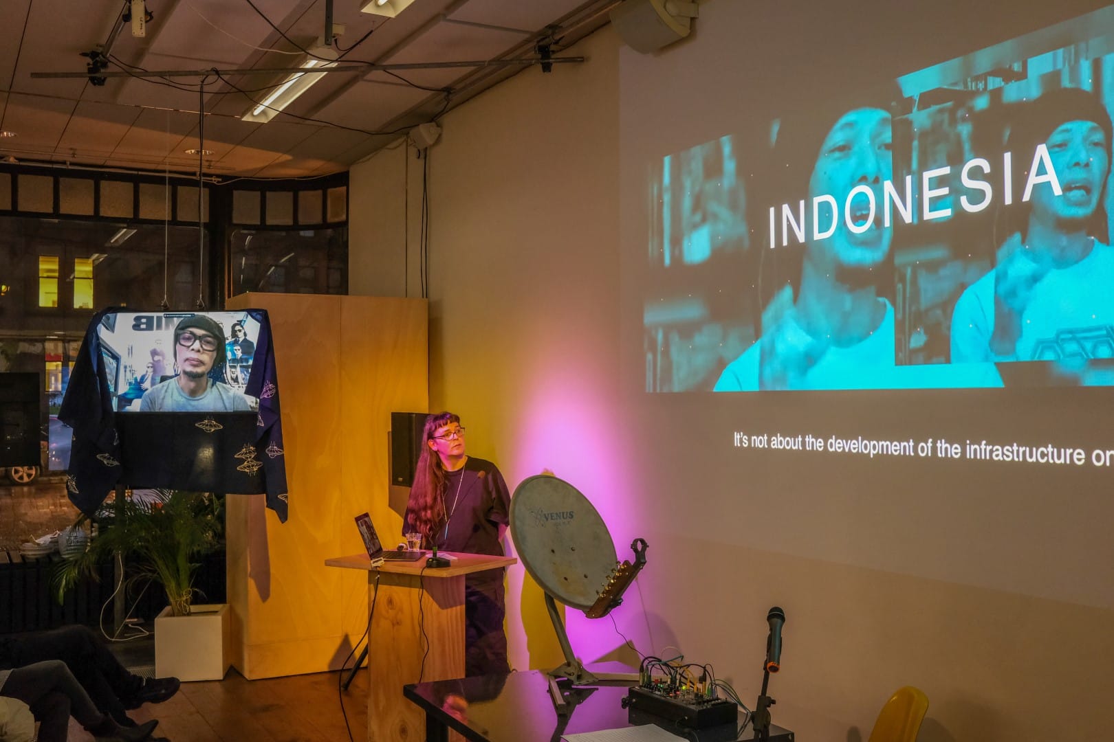 Adriana Knouf standing at a lectern, with Venzha Christ on a Zoom call on a TV hanging from the ceiling, draped by a dark blue batik fabric with UFO motifs on it.