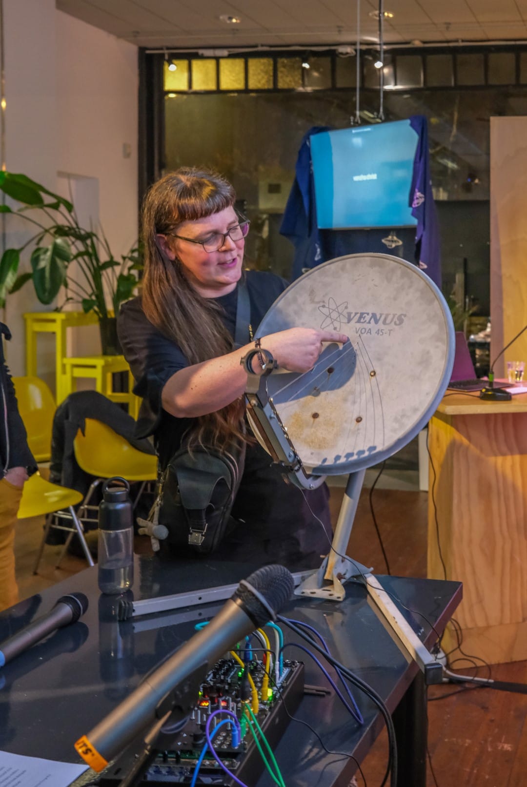 Adriana Knouf demonstrating her satellite dish instrument as it sits on the table. She is pointing to the deformation of the dish due to the tension of the strings.